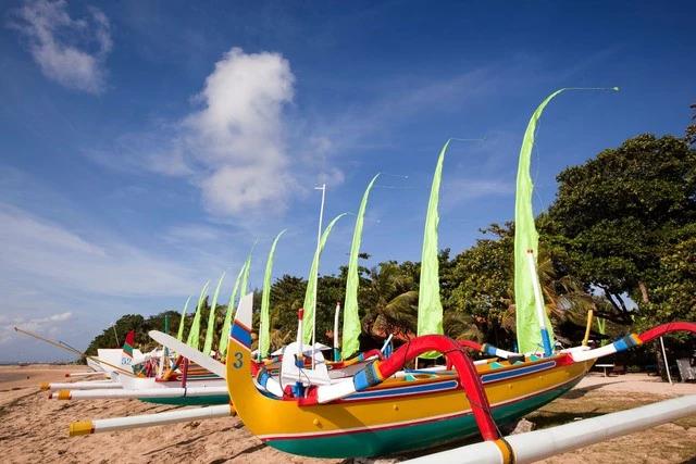 Traditional boats with tall sails and Bali-style flags at serene beach harbor.