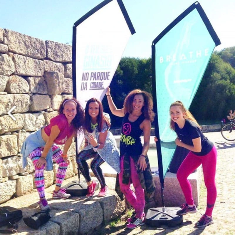 Four athletes demonstrating K banner deployment in park settings: white/teal fabric panels with water tank. Visible water tank base plates ensure stability on stone pavement during fitness events.