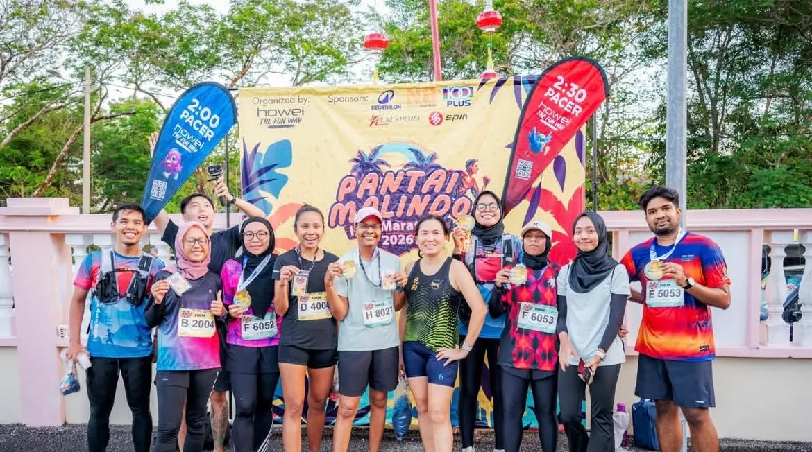Happy runners celebrate at the Pantai Malindo Marathon 2026, proudly displaying medals and bibs in front of the event banner.