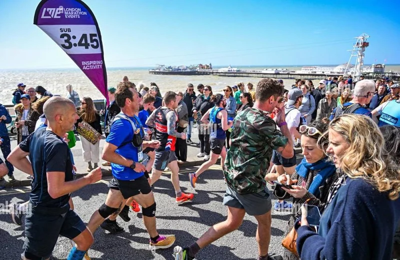 London Marathon pacer with purple 'SUB 3:45' teardrop flag on backpack, running by seafront with crowd