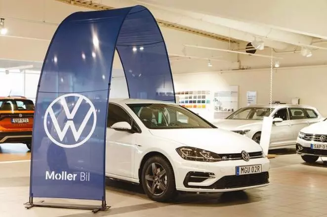 White Volkswagen sedan in dealership showroom with blue arch stand display, accompanied by orange/white vehicles under bright exhibition lighting.