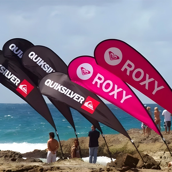 Beachside black and magenta flags with QUIKSILVER and ROXY logos, heart symbols, against ocean backdrop. Crowds on sand reflect coastal leisure vibe.