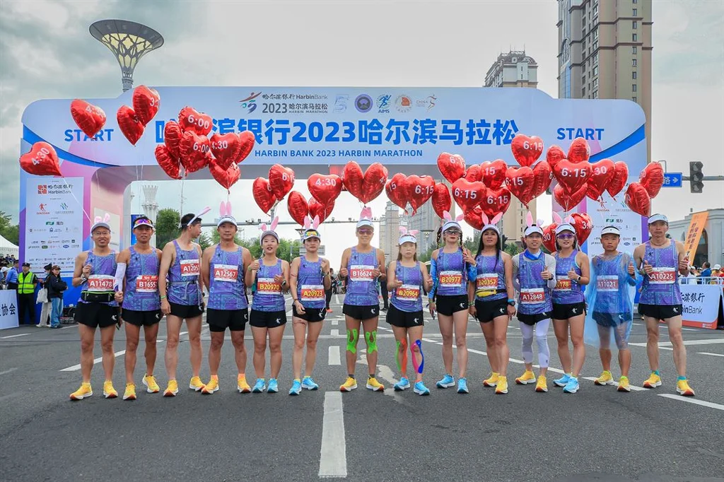 Harbin 2023 marathon start: runners in purple vests holding red '6:15' heart hung balloons under arch