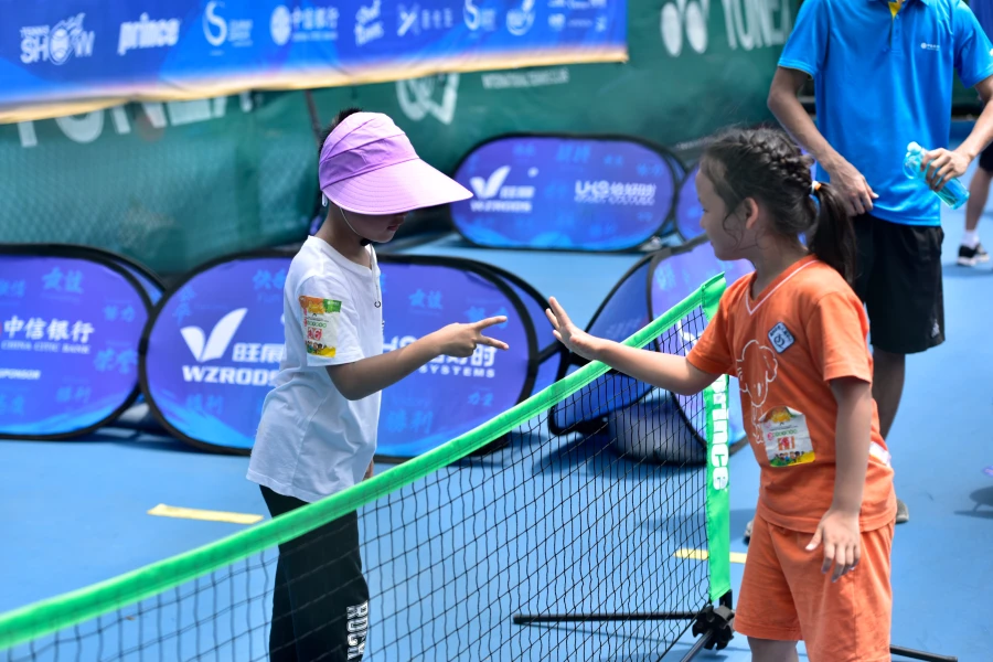 Children interacting at tennis net, blue sideline banners (WZRODS, UHS) behind, capturing event atmosphere.